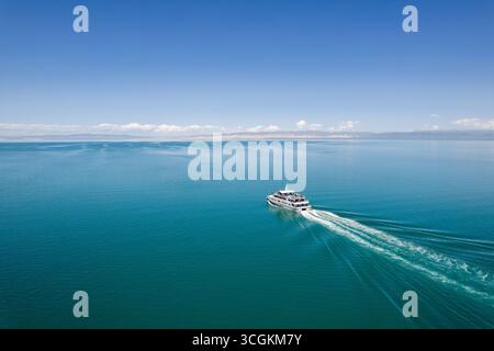 Ein großes Boot fährt unter hellem Himmel über das klare blaue Wasser des Qinghai Lake und bietet unvergessliche Reiseerlebnisse in der Nähe historischer Klöster. Stockfoto