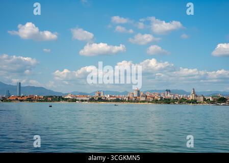 Die Stadt Xiamen in China zeigt ihre Skyline über dem Wasser. Städtische Gebäude stehen vor fernen Bergen, unter einem blauen Himmel voller weißer Wolken Stockfoto