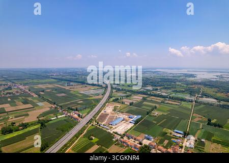 Unter klarem Himmel verstreute Landfelder zeigen eine Autobahn, die sich durch grünes Ackerland schlängelt, mit einem weit entfernten Gewässer. Stockfoto