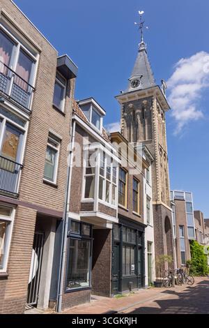 Eine enge Straße in Gouda, gesäumt von historischen Backsteingebäuden und einem prominenten Barbaratorenturm mit einer Wetterfahne vor blauem Himmel. Stockfoto