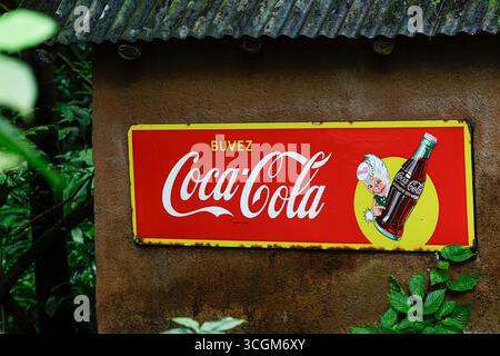Vintage Coca-Cola-Schild mit französischem Text auf einer rustikalen Wand, umgeben von Grün. Stockfoto