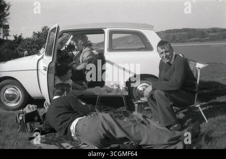 1960er Jahre, historisch, auf grasbewachsenem Land am See, eine Familie, die ein Picknick vor ihrem Auto aus der damaligen Zeit machte, ein Austin A40 Farina, hergestellt von der British Motor Corporation (BMC). Der dreitürige Kleinwagen wurde von 1958 bis 1967 produziert. Stockfoto