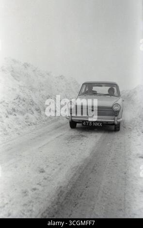 1960er Jahre, historische Vorderansicht eines dreitürigen Austin A40 Farina Motorwagens aus der Zeit auf einer schneebedeckten Strecke auf einer Tour durch den Kontinent. Dad fährt, Tochter sitzt neben ihm. Mit einem italienischen Design von Battista Farina und in Produktion zwischen 1958 und 1967 von der British Motor Corporation (BMC) war der Austin A40 der letzte der vielen Autos mit diesem Namen. Stockfoto