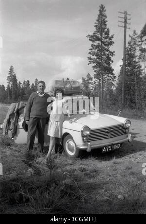1960er Jahre, historisch, ein dreitüriger Austin A40 Farina-Motorwagen der Epoche auf einer Tour durch den Kontinent. Das Gepäck auf dem Dachträger, ein Paar steht im Auto für ein Foto, das am Straßenrand neben einem Wald geparkt ist. Der A40 wurde zwischen 1958 und 1967 in Produktion genommen, mit italienischem Design von Battista Farina. Der Austin A40 wurde von der British Motor Corporation (BMC) hergestellt und war der letzte der Wagen mit diesem Namen. Stockfoto