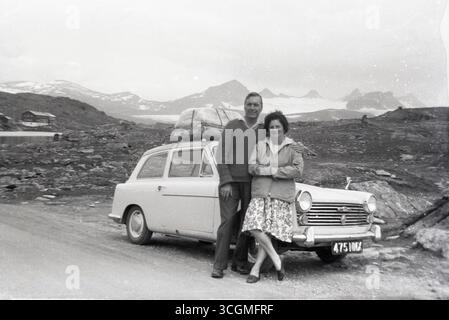 1960er Jahre, historisch, ein dreitüriger Austin A40 Farina-Motorwagen der Epoche auf einer Tour durch den Kontinent. Das Gepäck auf dem Dachträger, ein Paar steht im Auto für ein Foto, das am Straßenrand oben geparkt ist. Der A40 wurde zwischen 1958 und 1967 in Produktion genommen, mit italienischem Design von Battista Farina. Der Austin A40 wurde von der British Motor Corporation (BMC) hergestellt und war der letzte der Wagen mit diesem Namen. Stockfoto