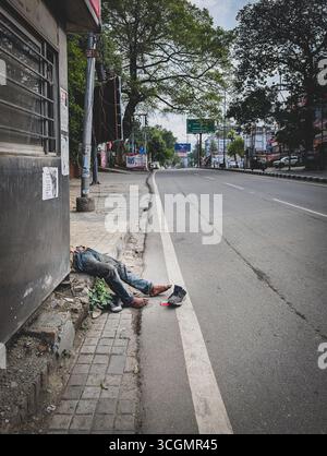 27. Mai 2025 dehradun City Indien. Betrunkener Obdachloser, der auf einem ruhigen Bürgersteig neben einer leeren Straße liegt, die von Bäumen und Gebäuden gesäumt ist Stockfoto
