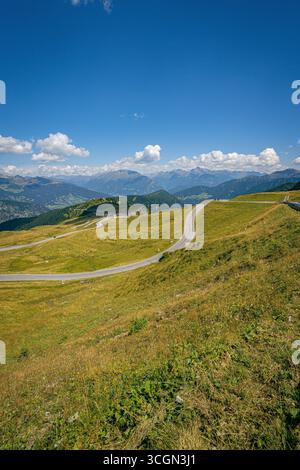 Gewundene Alpenstraße über grasbewachsene Hügel mit Panoramablick auf entfernte Bergketten der Dolomiten unter klarem blauen Sommerhimmel Stockfoto