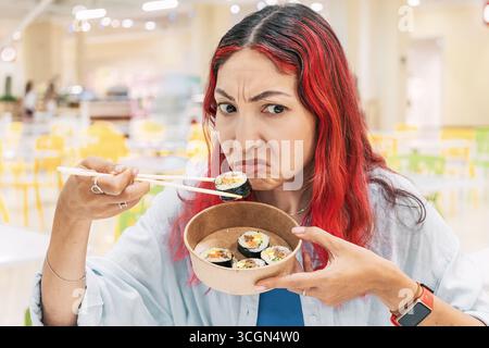 Eine junge Frau, die in einem belebten Speisesaal Kimbap oder Sushi mit Essstäbchen isst und ihre Unzufriedenheit über verdorbenes Essen zum Ausdruck bringt Stockfoto
