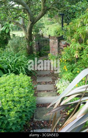 Ein ruhiger Pfad aus Steintreppen schlängelt sich durch lebendiges Grün und führt zu einem bezaubernden Tor mit üppigen Pflanzen und bunten Blumen, die die Gegend umgeben. Stockfoto