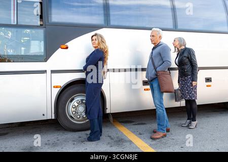 Drei Personen stehen an einer Bushaltestelle neben einem geparkten Reisebus an einem sonnigen Tag in entspannter Kleidung auf ihren Bus Stockfoto