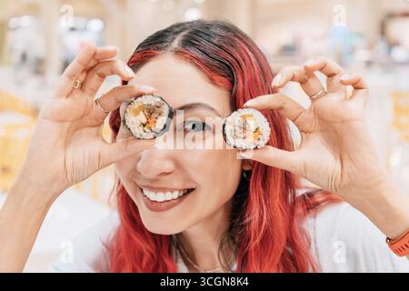 Eine verspielte junge Frau mit leuchtend roten Haaren genießt einen köstlichen Kimbap-Snack, der ihrem Essen einen Hauch von Spaß verleiht Stockfoto