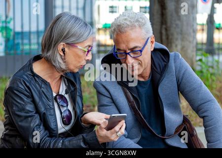 Ein reifes Paar Pendler kommentiert fröhlich ihre Smartphones, während sie draußen sitzen Stockfoto