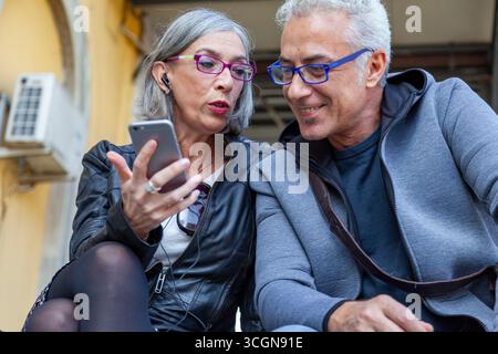 Niedrigwinkelporträt eines lächelnden, Reifen Paares männlicher und weiblicher Pendler, die Smartphones benutzen, während sie draußen an einem Bahnhof sitzen Stockfoto