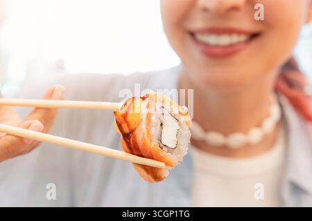Lächelnde Frau, die hölzerne Essstäbchen mit Sushi-Brötchen hält und kurz davor ist, es zu essen und japanische Küche zu genießen Stockfoto