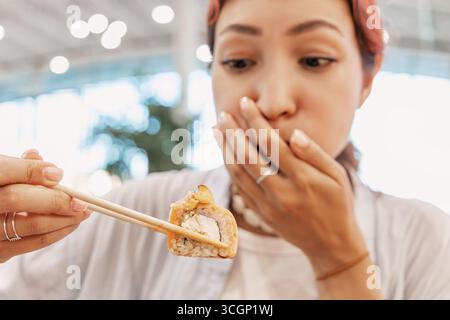 Die junge Frau hält eine Sushi-Rolle mit Stäbchen und bedeckt ihren Mund und drückt Ekel aus Stockfoto