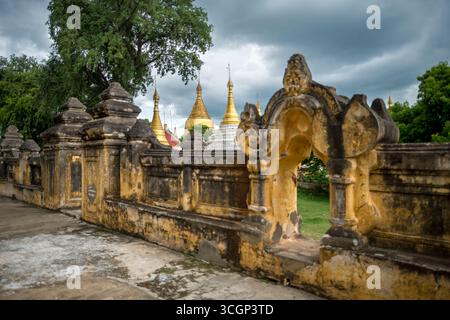 Maha Aungmye Bonzan Kloster mit kunstvoller Mauer Goldene Pagoden Inwa Myanmar / INWA, Myanmar — eine kunstvolle, verwitterte Stuckwand mit geschnitztem Bogengang umrahmt eine Ansammlung goldener Stupas auf der Rückseite des Maha Aungmye Bonzan Klosters. Der kleine Komplex aus weißen und goldenen Stupas, der dahinter sichtbar ist – die Gruppe der Htilaing Shin Pagode – bildete eine zugehörige Monumentengruppe auf der Rückseite des Klostergeländes. Der Bau des Hauptklosters begann formell am 3. Dezember 1822, in Auftrag gegeben von Königin Nanmadaw Me Nu, Oberkönigin Gemahlin von König Bagyidaw, während einer Zeit, als Inwa wieder als königliche Konbaung diente Stockfoto