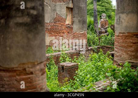 Yadana Hsemee Pagode Komplex Buddha Statue Ruinen Inwa Myanmar // AMARAPURA, Myanmar (Burma) – Eine verwitterte Buddha Statue befindet sich inmitten der Backsteinruinen des Yadana Hsemee Pagode Komplexes in Inwa (auch bekannt als Ava). Diese antike buddhistische Stätte, die von bröckelnden Ziegelmauern und Steinsäulen geprägt ist, ist heute weitgehend mit grünem Laub bewachsen. Der Yadana Hsemee-Komplex ist ein wichtiger Teil des historischen archäologischen Gebiets Inwa, das vom 14. Bis zum 19. Jahrhundert als königliche Hauptstadt für mehrere burmesische Königreiche diente. Diese atmosphärischen Ruinen bieten einen ergreifenden Einblick in M Stockfoto