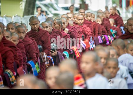 Mahagandayon Kloster Novizen Mittagsprozession Mandalay Myanmar / AMARAPURA, Myanmar — buddhistische Novizen, bekannt als Samanera, tragen ihre Almosenschalen durch die tägliche Mittagsprozession im Mahagandayon Kloster. Sie bewegen sich in geordnetem Schweigen durch das Gelände und erhalten Nahrung von Laiengeweihten – eine Praxis, die das Vertrauen der klösterlichen Gemeinschaft und ihre wechselseitige Beziehung zu der Laiengemeinschaft symbolisiert, der sie dient. Mahagandayon ist eine der größten und bekanntesten Klosterhochschulen in Myanmar, die Heimat von über 1.000 Mönchen und Novizen in Amarapura, etwa 11 km (7 Meilen) so Stockfoto