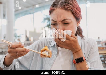 Die junge Frau hält eine Sushi-Rolle mit Essstäbchen und bedeckt ihre Nase mit einer Serviette und drückt Ekel aus Stockfoto
