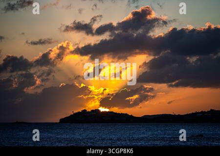 Sonnenuntergang über St. Thomas vom Gallows Point Cruz Bay St. John USVI // St. JOHN, USVI — die Sonne geht hinter St. Thomas in der Ferne unter, vom Gallows Point in Cruz Bay auf St. John auf den U.S. Jungferninseln. Der Himmel ist voller dramatischer Wolken, einige davon beleuchtet durch das goldene Licht der untergehenden Sonne. Die Silhouette der Insel St. Thomas ist am Horizont sichtbar, mit Gebäuden und Vegetation, die vor dem pulsierenden Himmel umrahmt werden. Das Wasser des Karibischen Meeres ist im Vordergrund dunkelblau und spiegelt die Farben des Sonnenuntergangs wider. Gallows Point ist ein beliebter Aussichtspunkt auf St. John, bekannt Stockfoto