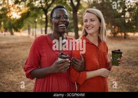 Zwei fröhliche Multi-ethnische Freunde lachen und spazieren in einem Park, während sie wiederverwendbare Kaffeetassen halten Stockfoto