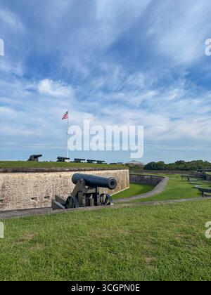 Eine Reihe historischer Kanonen entlang der grasbewachsenen Stadtmauer des Fort Macon State Park in North Carolina, über der die amerikanische Flagge stolziert weht. Stockfoto