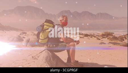 Sitzender Wanderer mittleren Alters mit roter Maske und Cargo Shorts am Sandstrand, mit grünem Rucksack Stockfoto