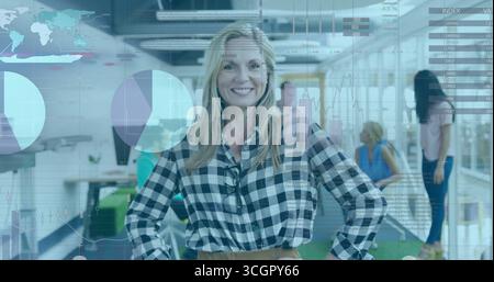 Blonde Frau in kariertem Hemd, die mit Händen auf Hüften im Großraumbüro steht, mit Datendiagrammen Stockfoto