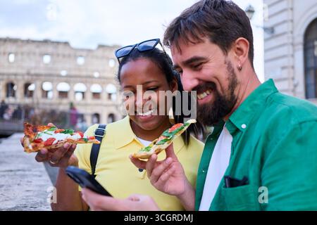 Glückliches Paar, das telefoniert und draußen Pizza vor dem kolosseum in Rom isst. Stockfoto