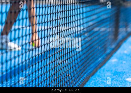 Nahaufnahme eines Padelnetzes mit einem unscharfen Spieler, der auf einem blauen Padelfeld läuft Stockfoto