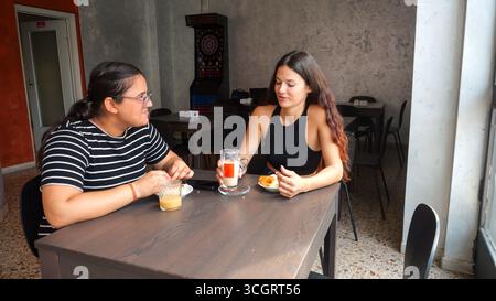 Zwei junge Freundinnen unterhalten sich tagsüber zwanglos und genießen Getränke und Snacks an einem Tisch in einer entspannten Terrasse im Freien Stockfoto