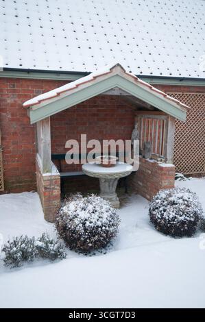 Ein bezaubernder Gartenpavillon mit schneebedeckten Steintischen und umliegendem Grün schafft eine friedliche Winteratmosphäre. Stockfoto