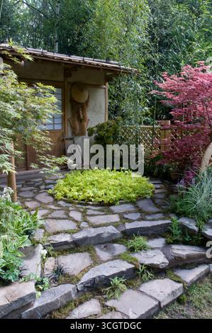 Der Garten verfügt über einen Steinweg mit üppigem Grün, der zu einem malerischen hölzernen Häuschen führt, eingebettet zwischen bunten Pflanzen. Stockfoto