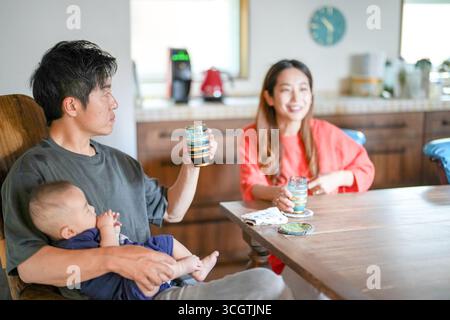 Ein japanischer Vater im Alter von 30 Jahren hält ein drei Monate altes Baby, während er an einem mediterranen Esstisch sitzt und Kaffee und Gespräche mit Th teilt Stockfoto