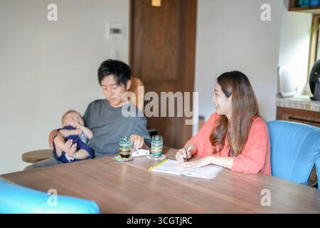 Ein Mann sitzt an einem Tisch mit einem Baby in den Armen. Eine Frau sitzt ihm gegenüber und schreibt in ein Notizbuch. Die Szene ist intim und friedlich, mit dem Mann A Stockfoto