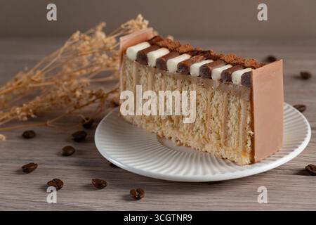 Nahaufnahme einer geschichteten Kaffeekuchenscheibe mit Schokolade und Sahne auf einem weißen Teller, umgeben von Kaffeebohnen und getrockneten Blumen Stockfoto