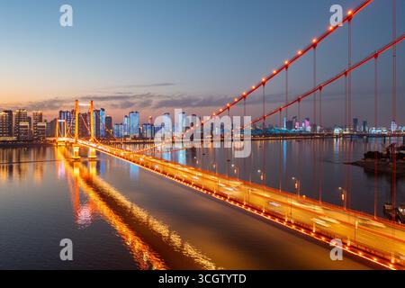 Die Wuhan-Brücke leuchtet hell über dem Wasser, während die Stadt in der Abenddämmerung erleuchtet. Stockfoto