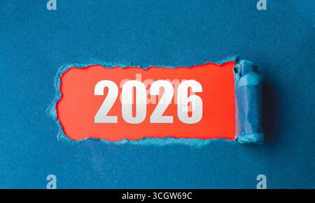 Eine große weiße 2026-Nummer auf rotem Hintergrund, die aus zerrissenem Papier auf blauem Hintergrund auftaucht. Eine Optik, die für das neue Jahr geeignet ist, Zukunftspläne Stockfoto