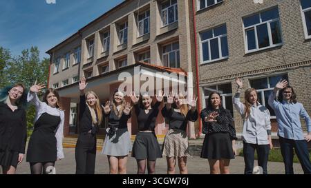 Glückliche Schüler winken vor dem Schulgebäude und feiern Einheit und Erfolge an einem sonnigen Tag Stockfoto