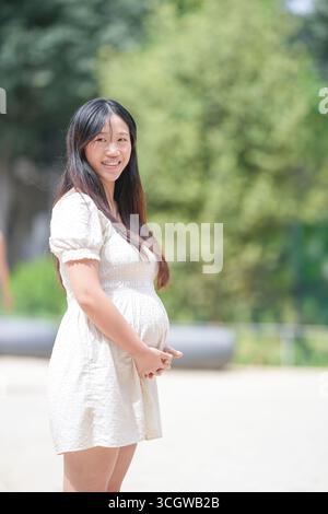 Eine Frau in einem weißen Kleid lächelt und hält ihren Bauch. Sie ist schwanger und genießt ihre Schwangerschaft Stockfoto