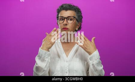 Ältere Frau mit grauem Haar und Brille entspannt sich vor einem leuchtend rosa Hintergrund, wobei sie sanft ihre Hände zur Selbstpflege und zur Linderung von Verspannungen in einem c Stockfoto