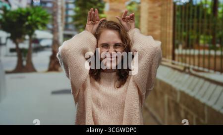 Frau macht lustige Gesichter in einer urbanen Straßenumgebung, die mit Brille und lockigem Haar im Freien in einer städtischen Umgebung mit Autom. Verspielt ausdrückt Stockfoto