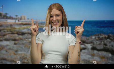 Die junge blonde Frau zeigt die Zeigefinger nach oben auf die felsige Küste an der sonnigen Küste; Optimismus Begeisterung Aufregung Freude. Stockfoto