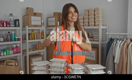 Die junge hispanische Frau meldet sich freiwillig in reflektierender Weste und macht eine Herzgeste in einem Spendenzentrum, das von Lebensmittelverpackungen und Kleiderregalen umgeben ist Stockfoto