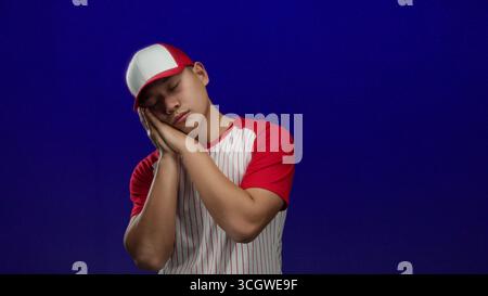 Junger chinesischer Mann in Baseballuniform mit roten und weißen Streifen, der schläfrig vor isoliertem blauem Hintergrund posiert, was auf einen müden Sportsportler hindeutet Stockfoto