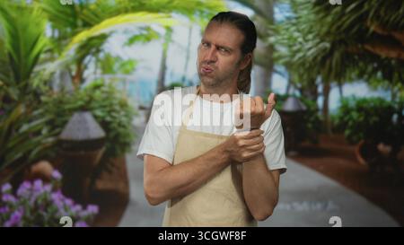 Mann trägt beige Schürzengrimaces, während er das Handgelenk auf einem von tropischem Grün gesäumten Parkweg hält; Unwohlsein. Stockfoto
