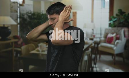 Der Mann bedeckt die Ohren mit beiden Händen zwischen gemütlichen Wohnzimmerstühlen und Lampe in einem Gebäudeinneren; Distress. Stockfoto