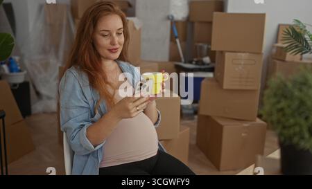 Schwangere Frau hält hellgelben Becher und Smartphone gegen sichtbaren Bauch im Gebäude; Mutterschaft Hoffnung. Stockfoto