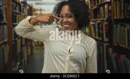 Frau mit lockigem Haar und Brille zeigt mit dem Finger auf Brille im alten Bibliotheksgebäude; Selbstvertrauen fokussiert. Stockfoto