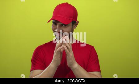 Hispanischer Kuriermann mit roter Kappe und Hemd drückt die Hände in einer steilen Geste im gelben Studio zusammen; listiger Strategieplan. Stockfoto
